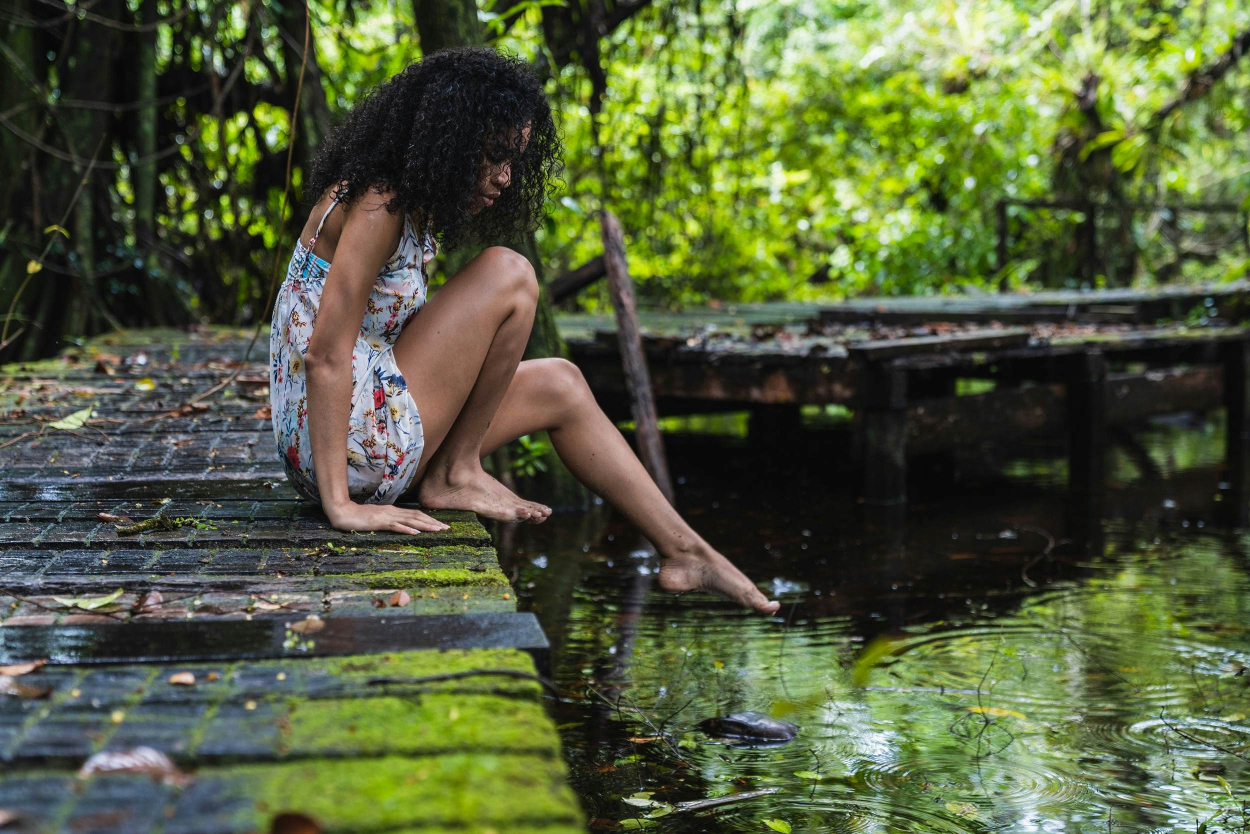 Woman with curly hair sitting on bridge stretches out leg towards water in green forest in Bocas del Toro Islands, Panama.
